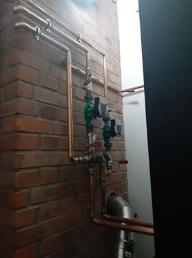 Miedziane rury instalacji wodnej i grzewczej zamontowane na ceglanej ścianie w Rawiczu, widoczne zawory i fragment komina.