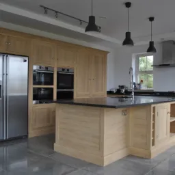 Jasna, drewniana kuchnia z wyspą i czarnym blatem, z widoczną lodówką ze stali nierdzewnej i dwoma piekarnikami w zabudowie.