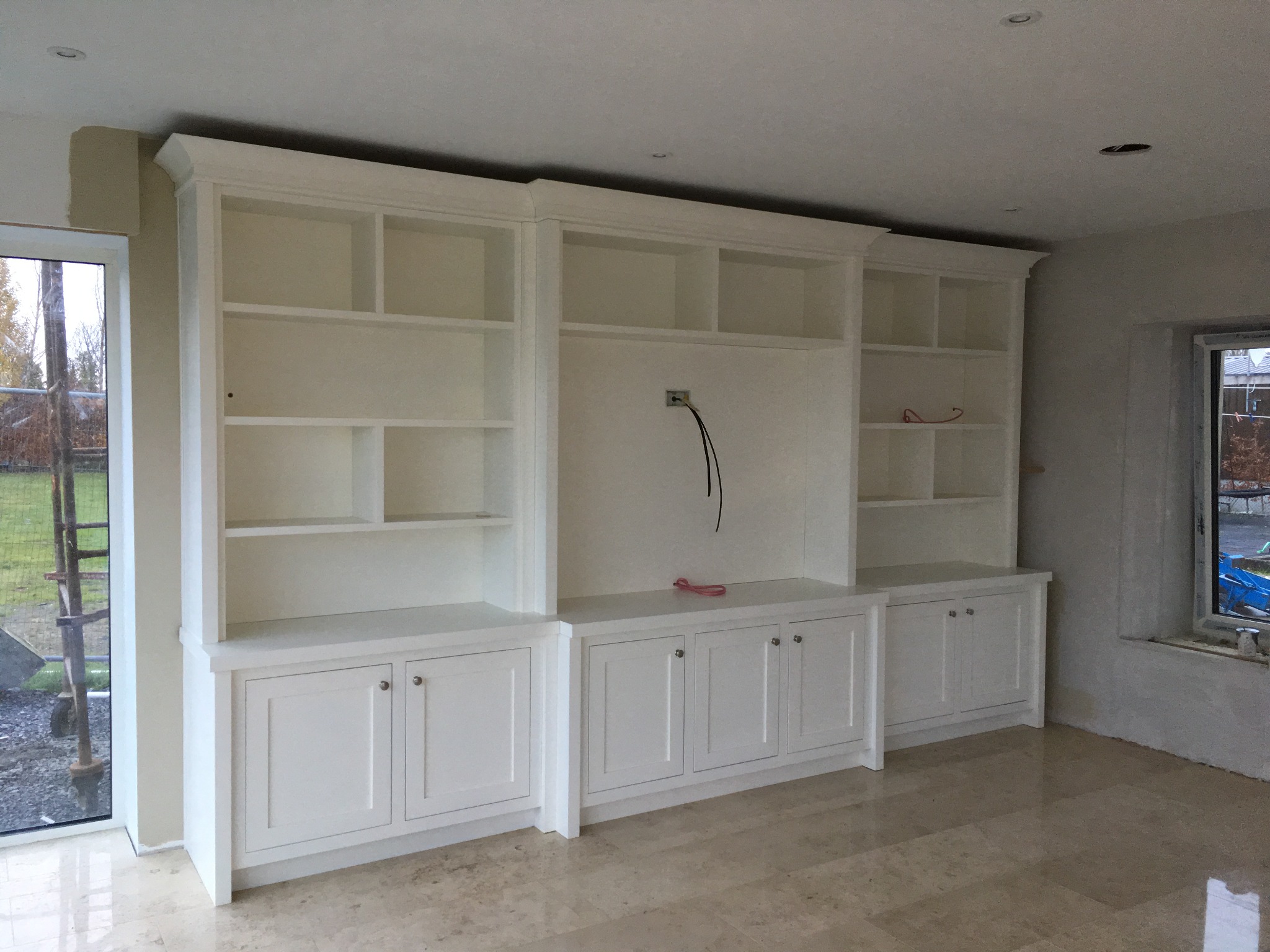 Biała, zabudowana szafa na wymiar z półkami i szafkami, widok na gniazdko elektryczne i kabel, marmurowa podłoga, okno z widokiem na zewnątrz.