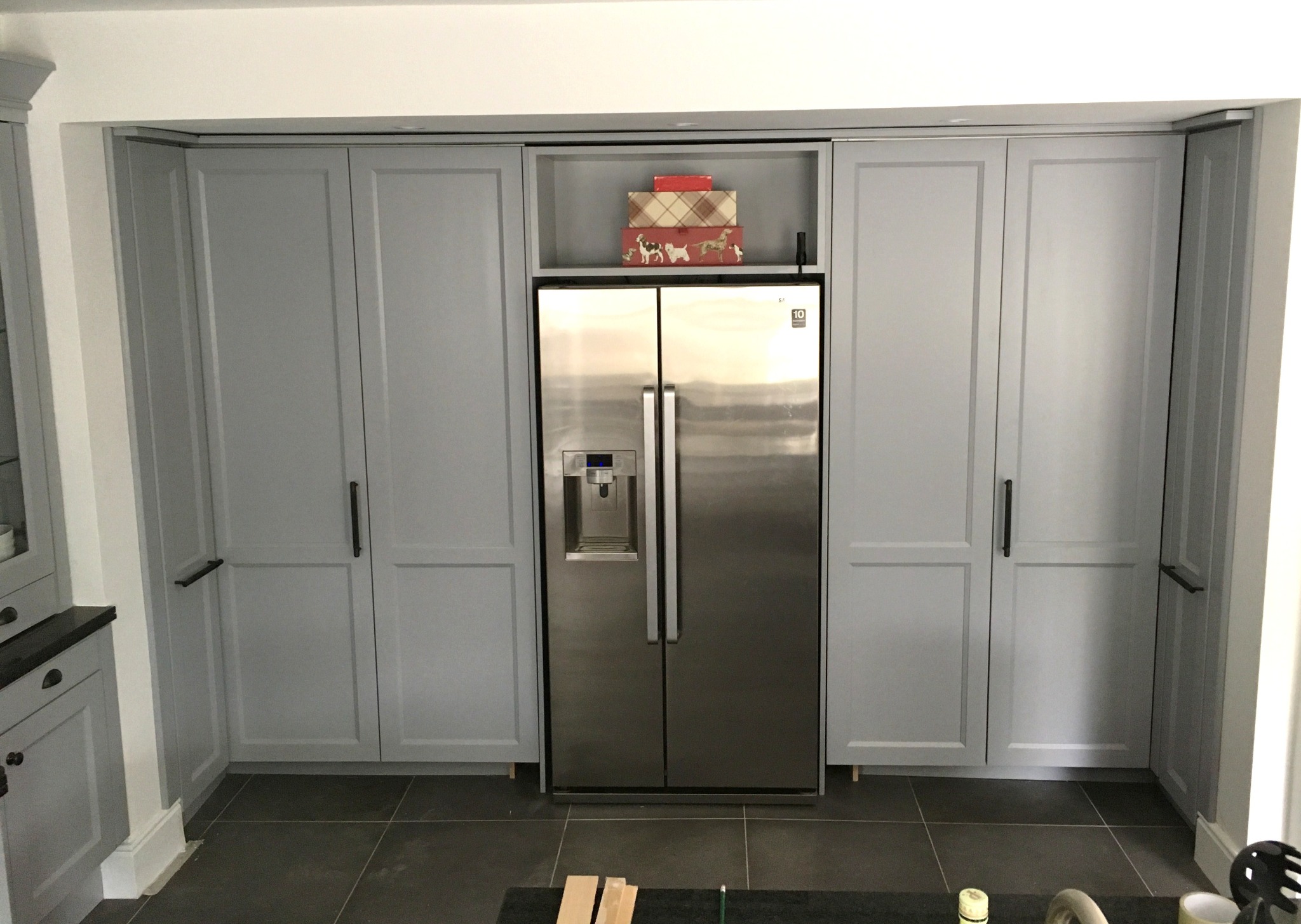 Szare szafy kuchenne na wymiar zabudowane wokół srebrnej lodówki dwudrzwiowej; na górze szafki dekoracyjne pudełka.