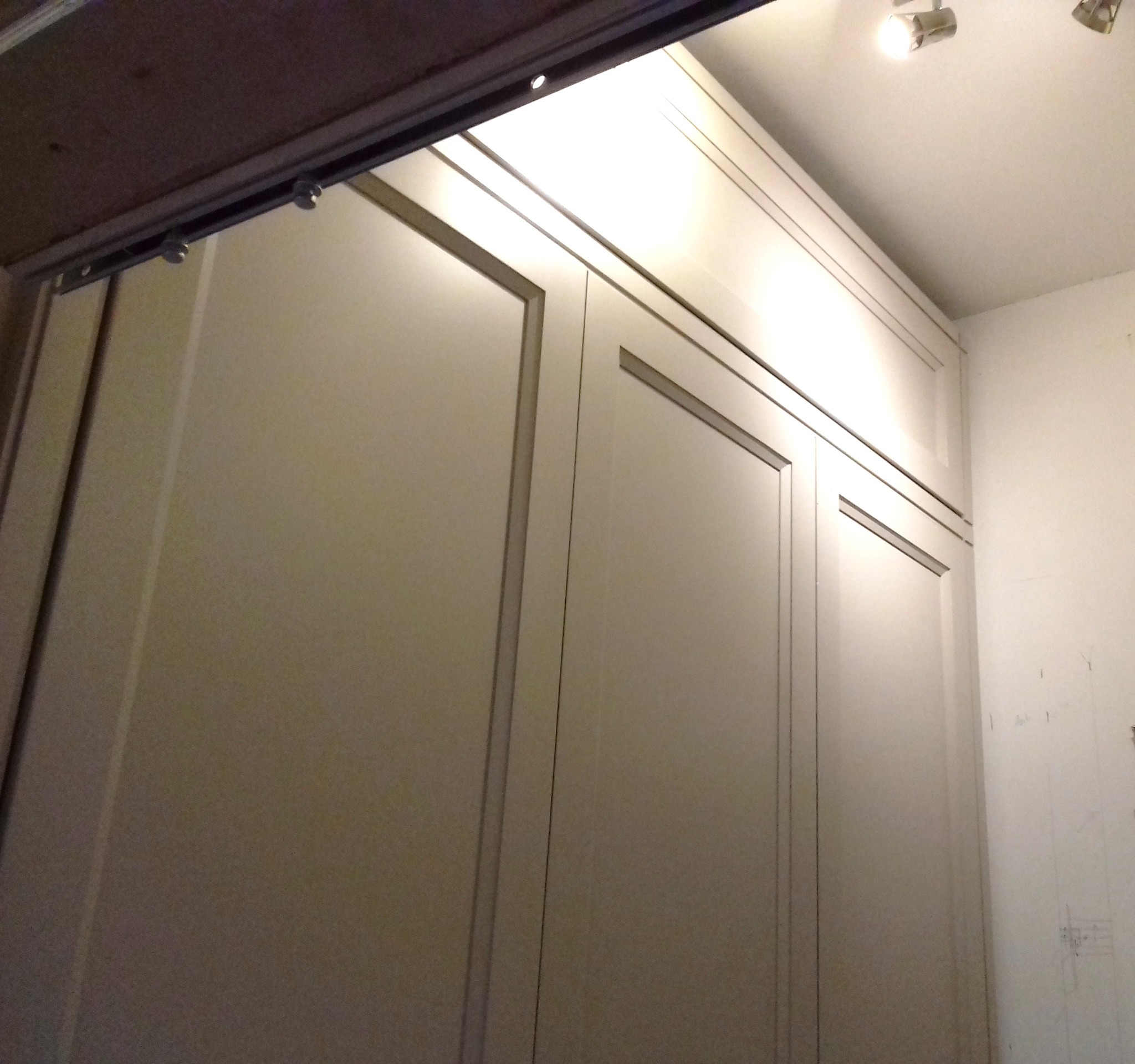 Beżowa szafa przesuwna z widocznymi prowadnicami i rolkami, zamontowana pod sufitem, oświetlona punktowo.