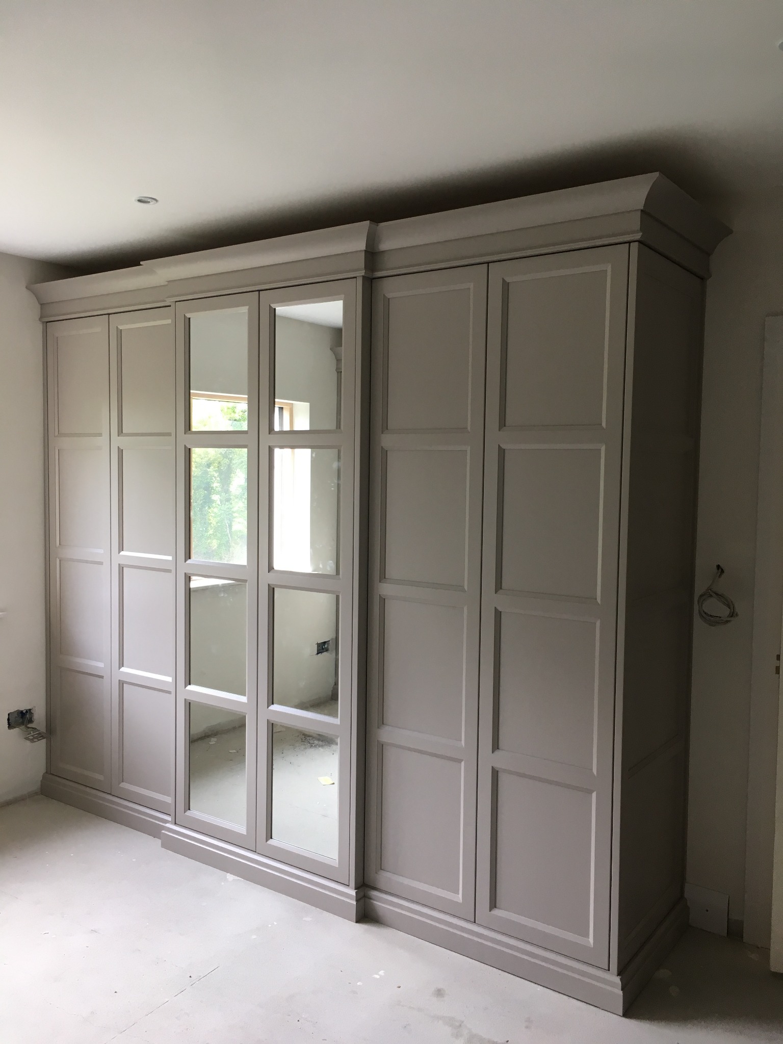 Duża, szara szafa wnękowa z lustrzanymi drzwiami, widoczne niedokończone wykończenie wnętrza, w tle okno z widokiem na zieleń.