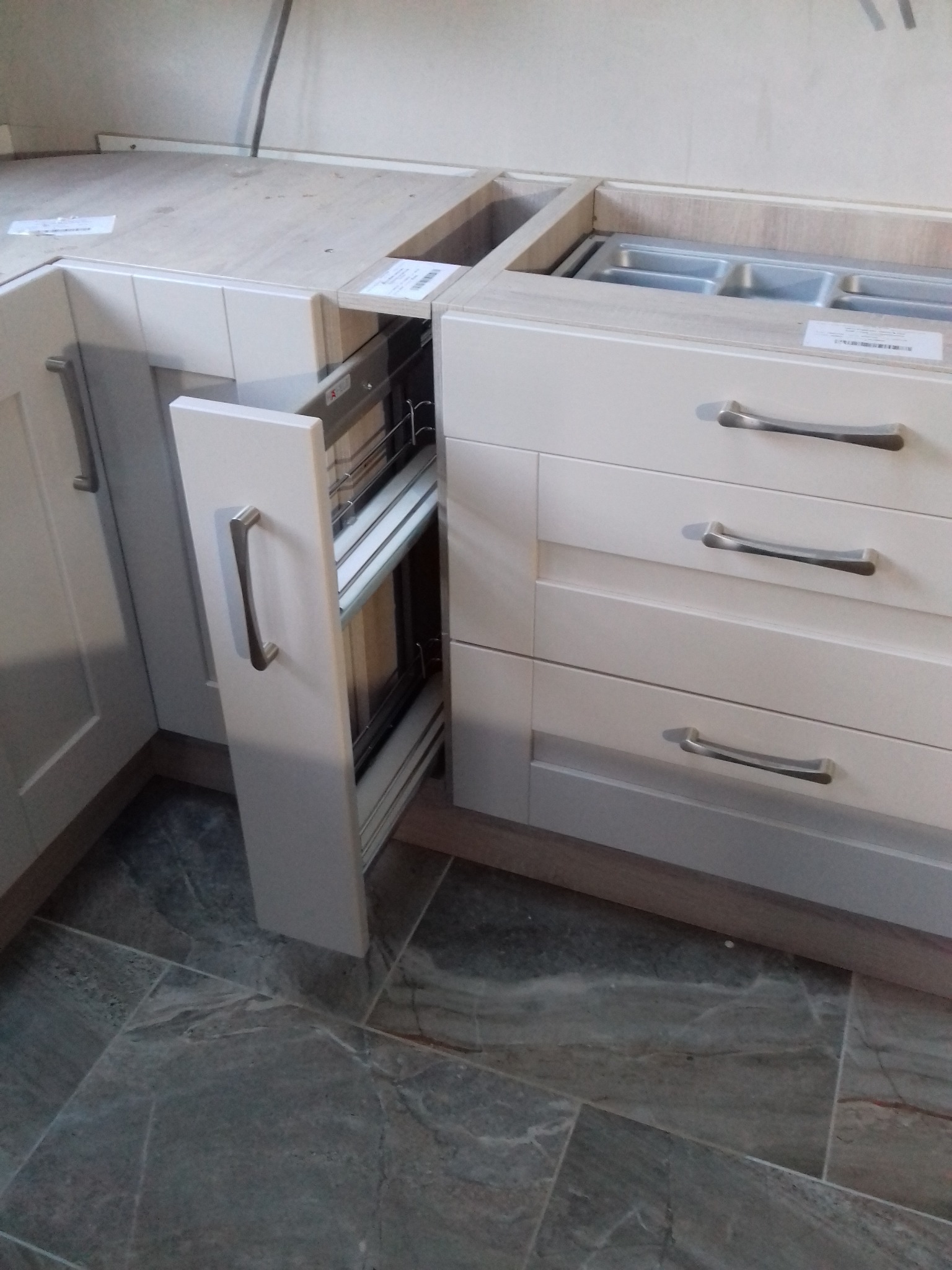 Szara szafka kuchenna z wysuwanym koszem cargo, widoczne trzy szuflady z metalowymi uchwytami i marmurowa podłoga.