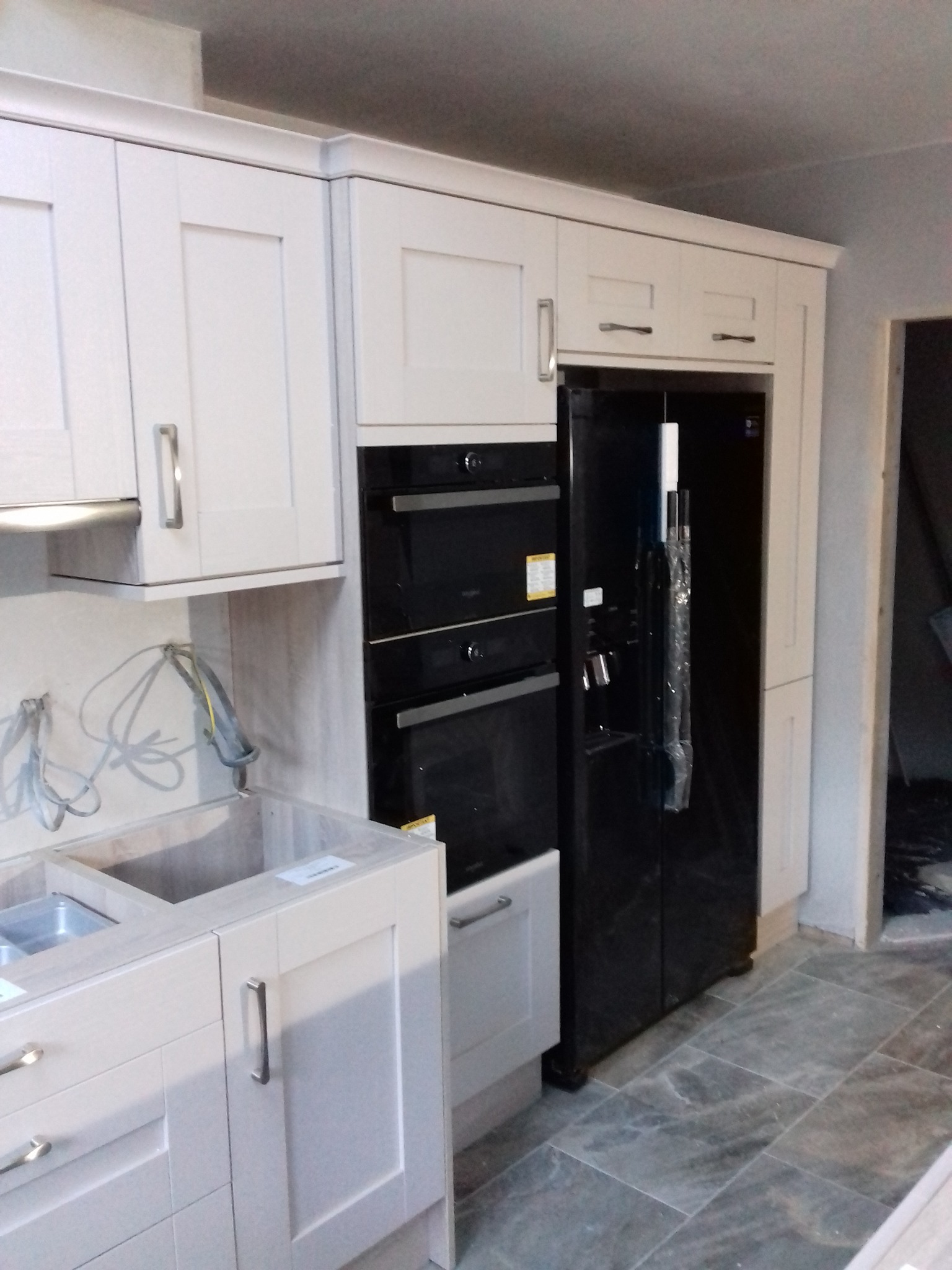 Montaż kuchni: białe szafki z frezowanymi frontami, wbudowany czarny piekarnik i lodówka side-by-side, widoczne przewody instalacji elektrycznej, szare płytki podłogowe.