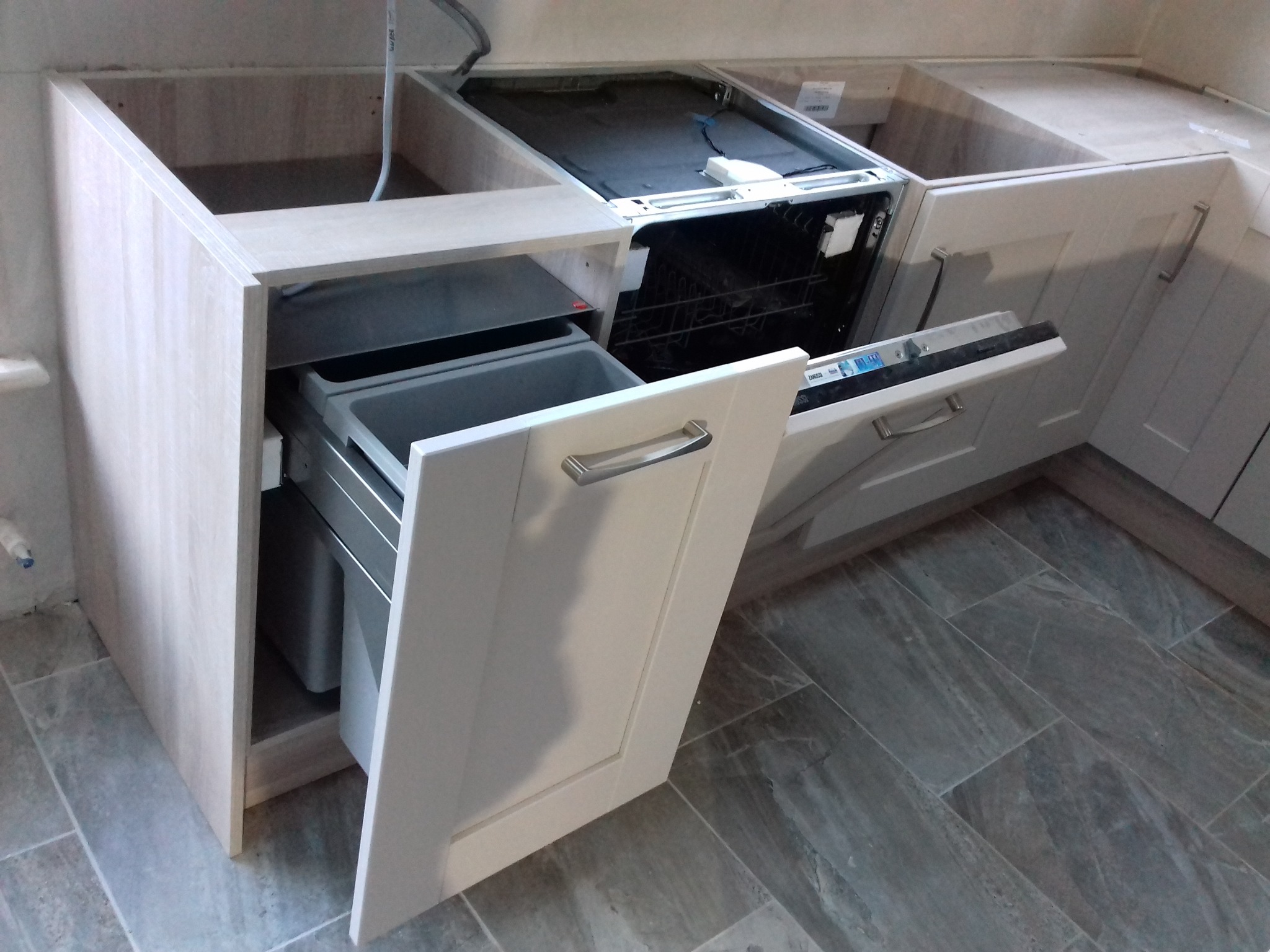 Montaż szafki kuchennej z wysuwanym koszem na śmieci i zmywarką w zabudowie, widoczne niedokończone wykończenie blatu.