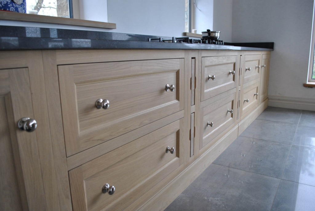 Jasne, drewniane szafki kuchenne z szarym blatem i okrągłymi, chromowanymi uchwytami, ustawione na szarej podłodze z płytek.