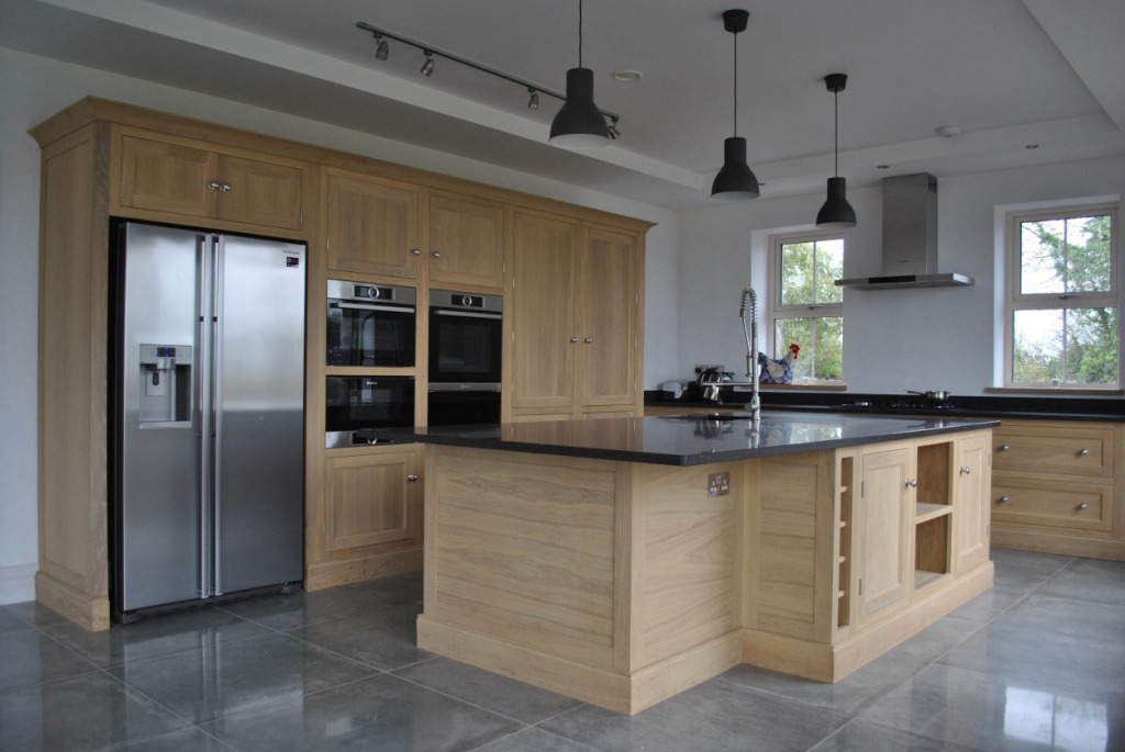 Jasna, drewniana kuchnia z wyspą i czarnym blatem, z widoczną lodówką ze stali nierdzewnej i dwoma piekarnikami w zabudowie.