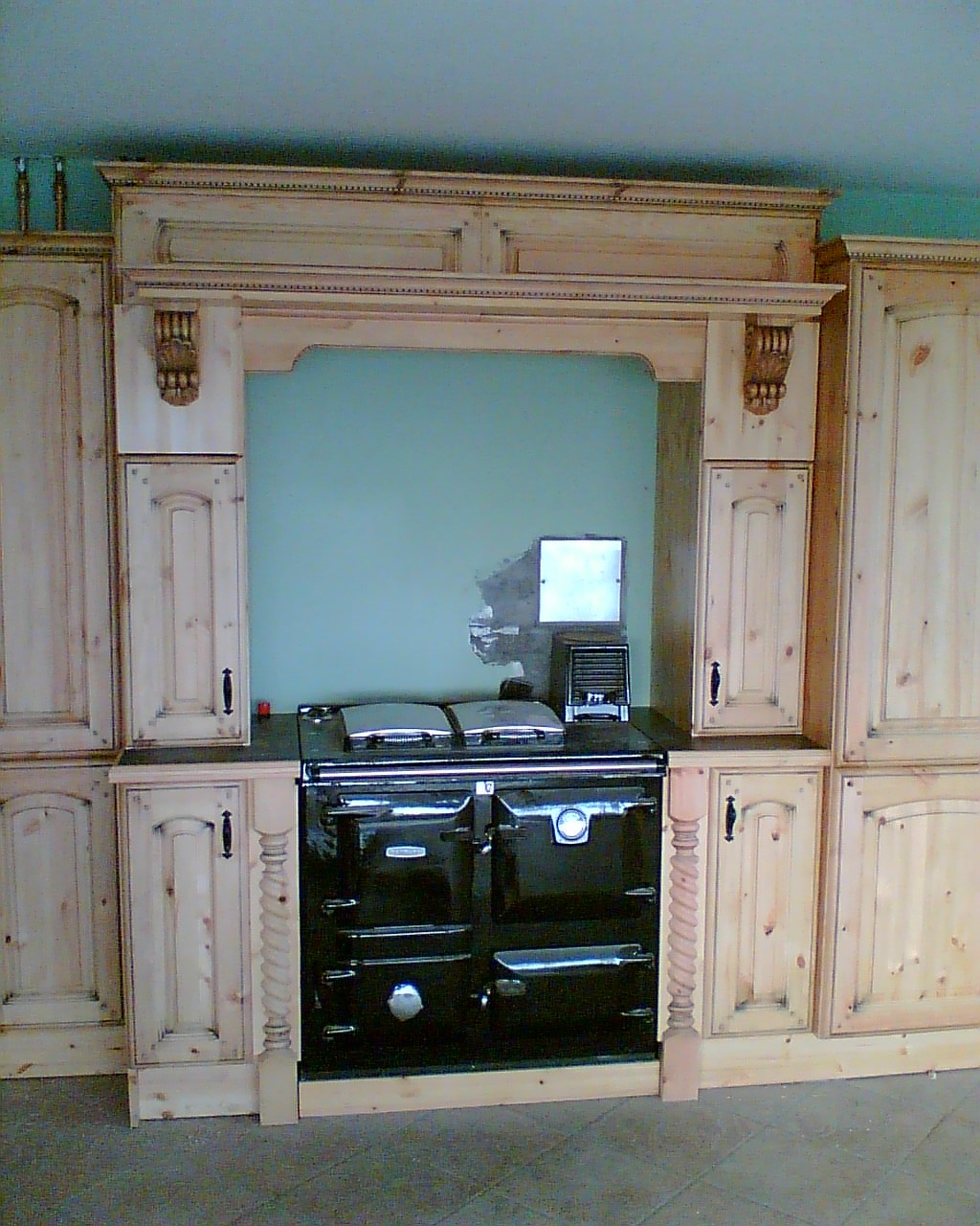 Czarny, emaliowany piec kuchenny z chromowanymi pokrywami, wkomponowany w zabudowę z surowego drewna z dekoracyjnymi elementami, na tle zielonej ściany.