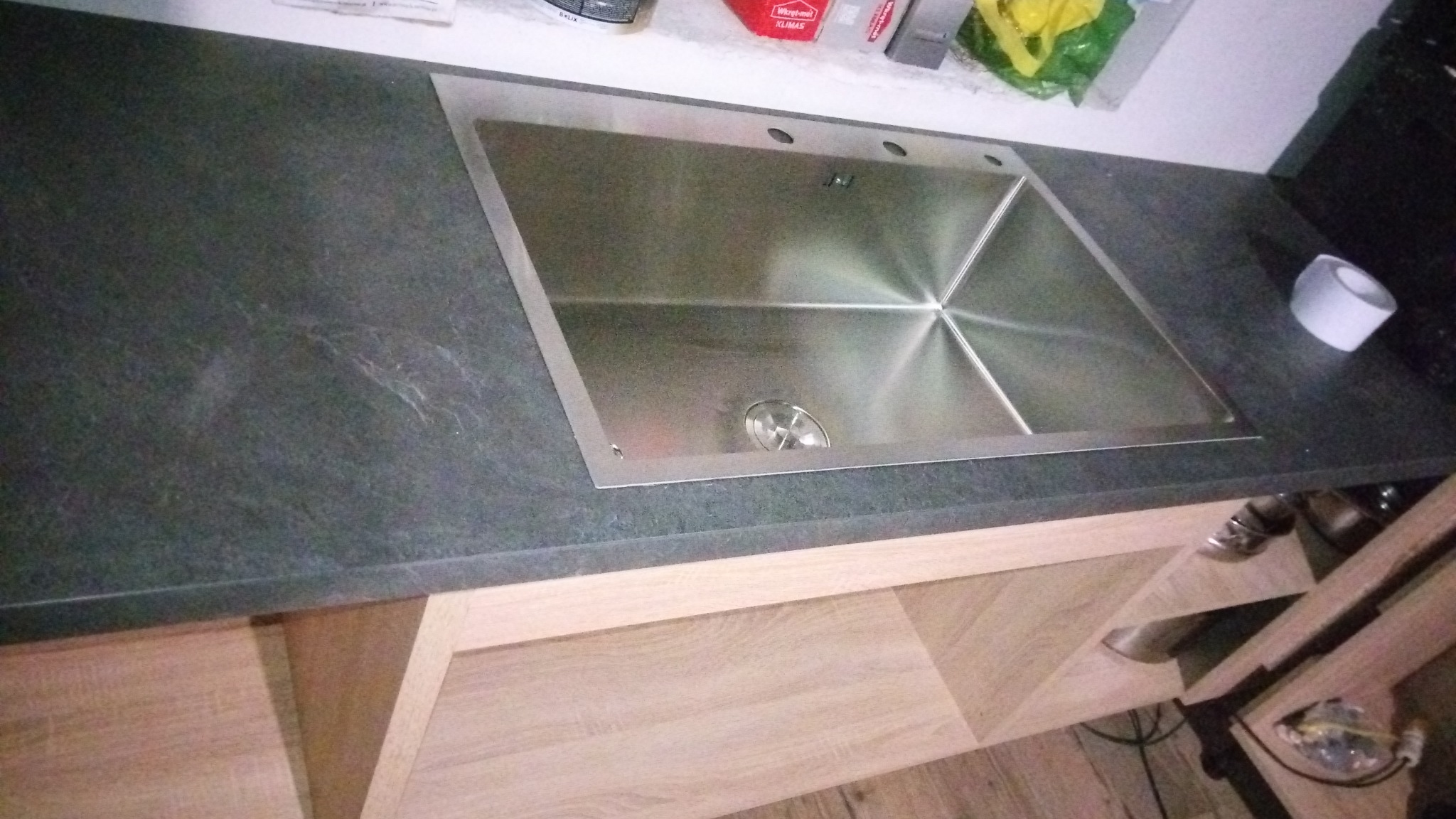 Montaż zlewozmywaka w blacie kuchennym o ciemnoszarej, nakrapianej powierzchni. Widoczna część szafki pod zlewem z jasnego drewna.