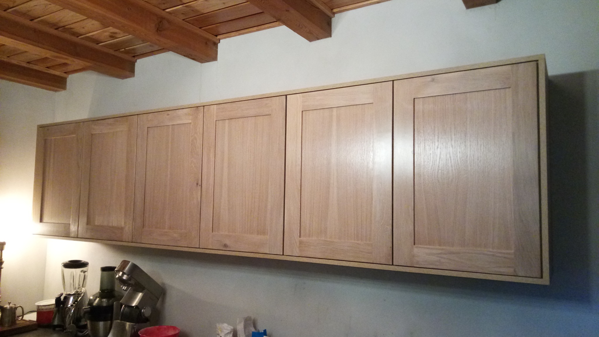 Wiszące szafki kuchenne z jasnego drewna, z widocznymi słojami, zamontowane pod drewnianym stropem z belkami, na blacie kuchennym widoczne urządzenia AGD.