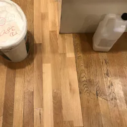 Fragment drewnianej podłogi, częściowo odnowionej, z widocznym wiadrem z produktem i kanistrem z płynem do pielęgnacji drewna, przy ścianie.