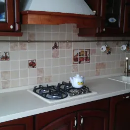 Kuchnia z ciemnymi drewnianymi szafkami, jasnym blatem i beżowymi płytkami z motywami owoców; na blacie biały czajnik obok płyty gazowej.