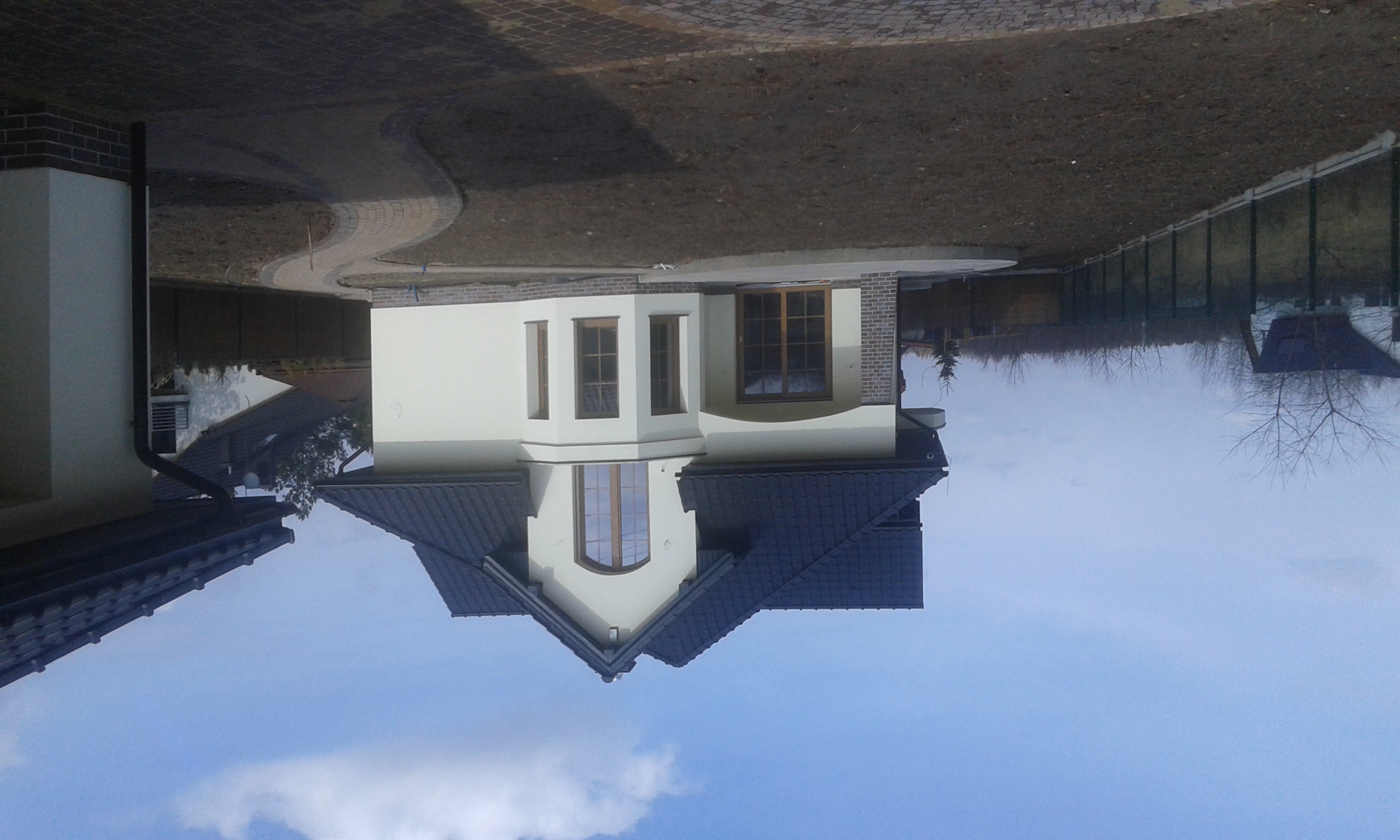 Nowoczesny dom jednorodzinny z jasną elewacją, ciemnym dachem i ceglanymi elementami wykończeniowymi, widoczny z perspektywy ogrodu w słoneczny dzień. Ścieżka z kostki brukowej prowadzi do domu.