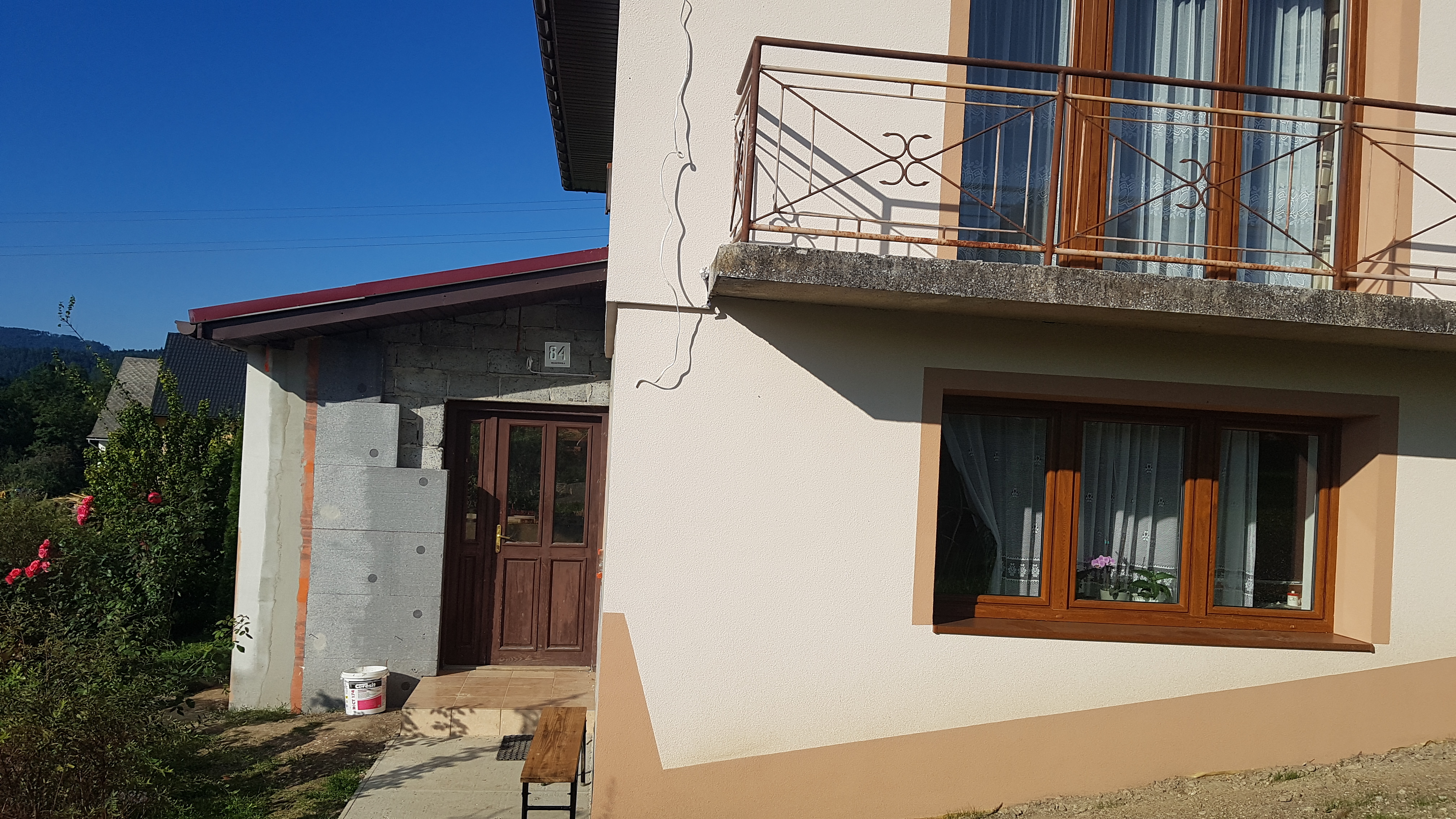 Fragment domu z ocieplonym styropianem fragmentem elewacji, drewnianymi oknami i balkonem z metalową balustradą.