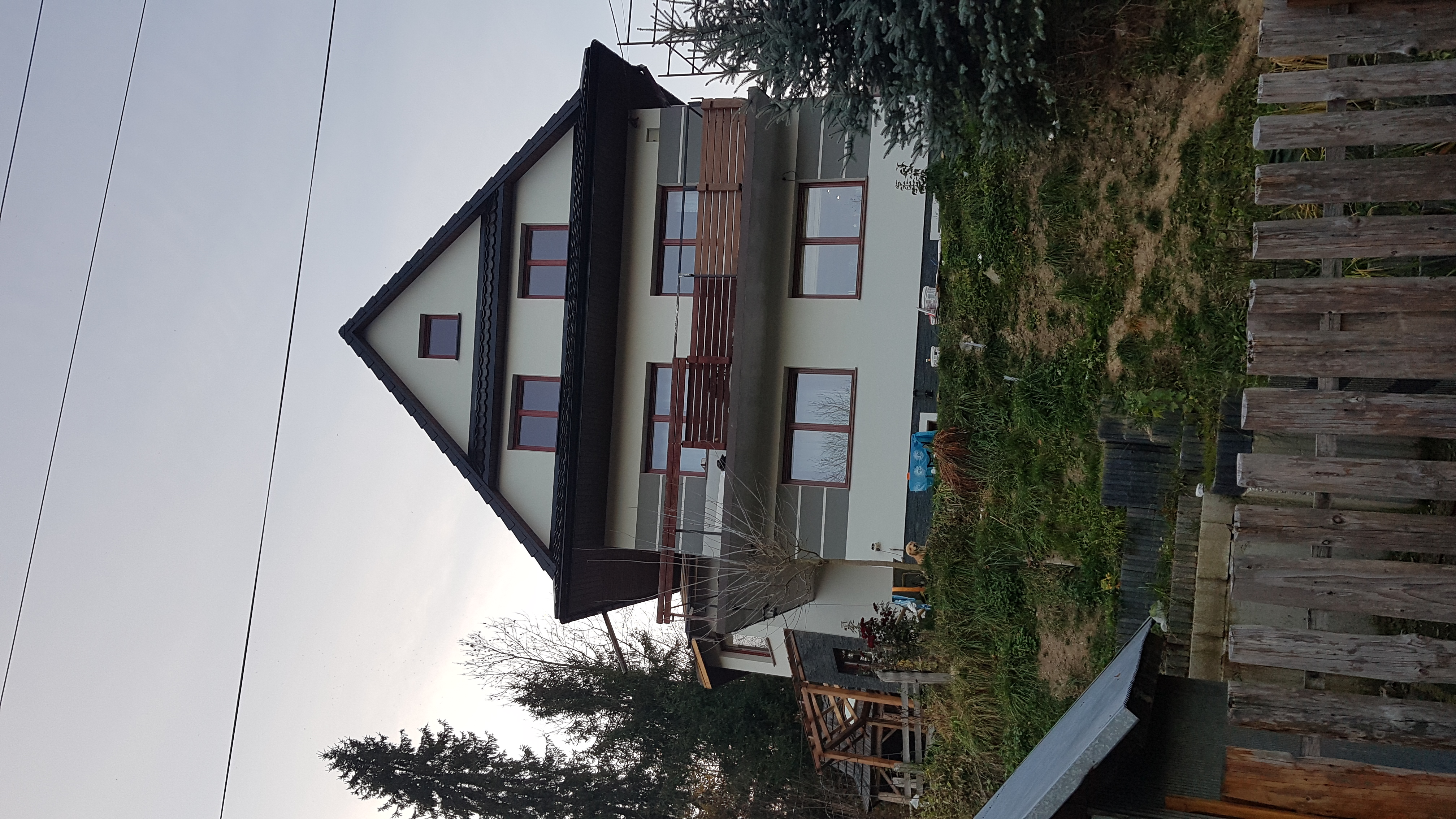 Dwupiętrowy dom z jasną elewacją i ciemnym dachem, widoczne dwa balkony z drewnianymi balustradami, otoczony zielenią i drzewami, ujęcie z dołu pod kątem.