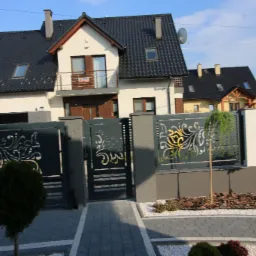 Nowoczesna posesja z ozdobnym ogrodzeniem metalowym w kolorze antracytowym z ażurowym wzorem, kostka brukowa i zadbana zieleń.