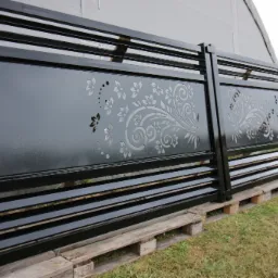 Czarny, metalowy segment ogrodzenia z laserowo wycinanym, floralnym wzorem, oparty na drewnianych paletach na trawie.