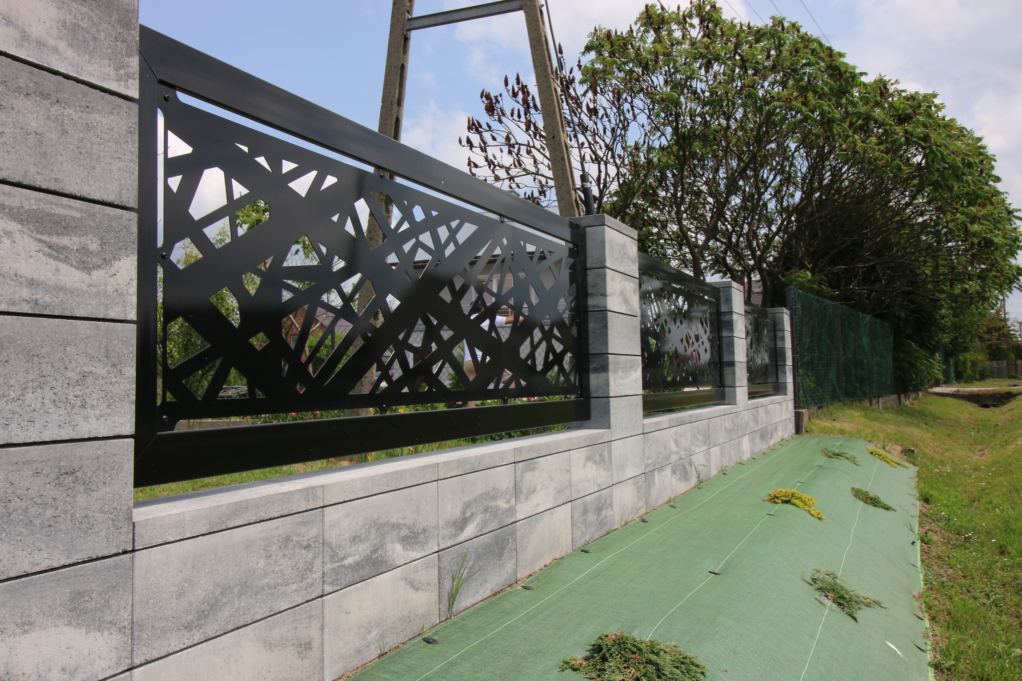 Nowoczesne ogrodzenie z czarnymi, ażurowymi panelami i szarymi, betonowymi słupkami, widok z boku na zieloną tkaninę ściółkującą i fragment krajobrazu.