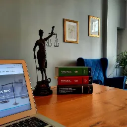 Biuro z drewnianym biurkiem, na którym stoi laptop z wyświetloną stroną internetową, statuetka Temidy, stos książek prawniczych (Prawo Cywilne, Prawo Karne, Czynności Procesowe), a w tle widoczny...