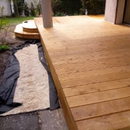 Nowo wybudowany taras z desek drewnianych, z widocznym podłożem przygotowanym pod dalszą część prac wykończeniowych, obok domu z dużym oknem.