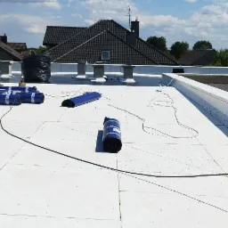 Biała powierzchnia dachu płaskiego z rozłożonymi rolkami niebieskiej membrany dachowej, czarnym workiem i odkurzaczem przemysłowym w trakcie prac. W tle zabudowa komina i domy jednorodzinne.