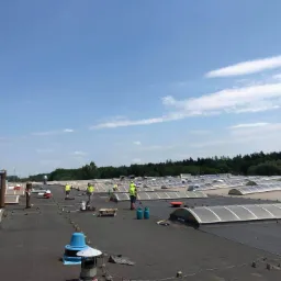 Prace na dachu hali: ekipa dekarzy w żółtych kamizelkach termicznie zgrzewa papę na płaskim dachu z świetlikami dachowymi, widoczne butle z gazem i narzędzia dekarskie.