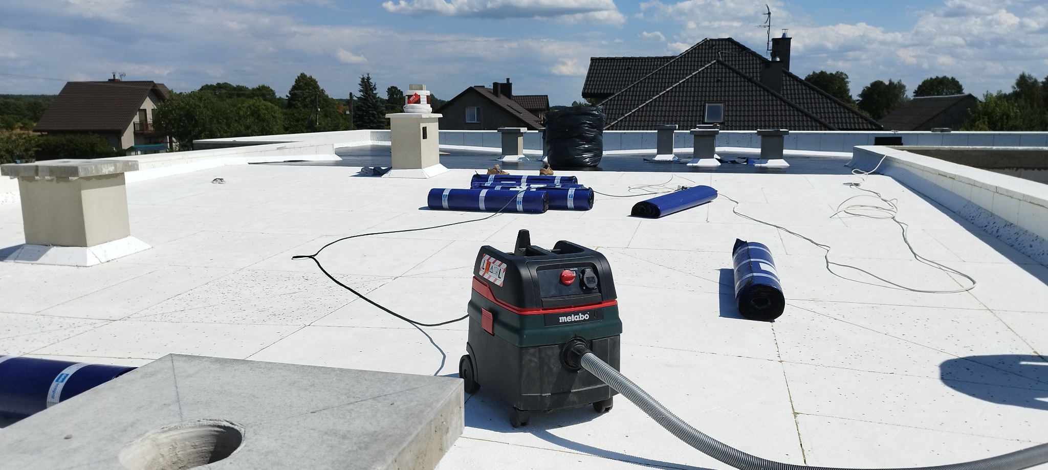 Biały dach płaski z widocznymi kominami, zrolowanymi membranami w kolorze niebieskim, czarnym workiem, odkurzaczem przemysłowym Metabo i betonowymi elementami wentylacyjnymi