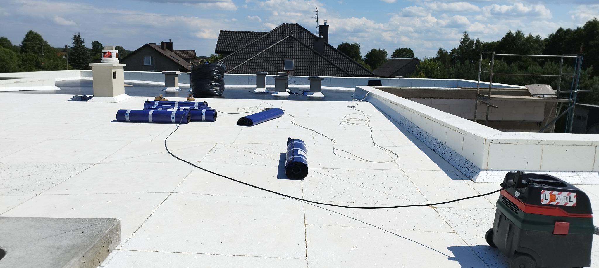 Biała powierzchnia dachu płaskiego z rozłożonymi rolkami niebieskiej membrany dachowej, czarnym workiem i odkurzaczem przemysłowym w trakcie prac. W tle zabudowa komina i domy jednorodzinne.