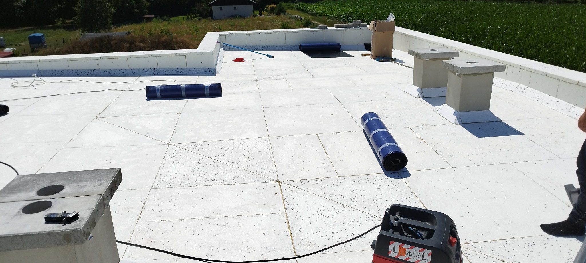 Płaski dach pokryty jasnymi płytami z widocznymi rolkami materiału izolacyjnego, narzędziami i elementami wentylacyjnymi w słoneczny dzień.