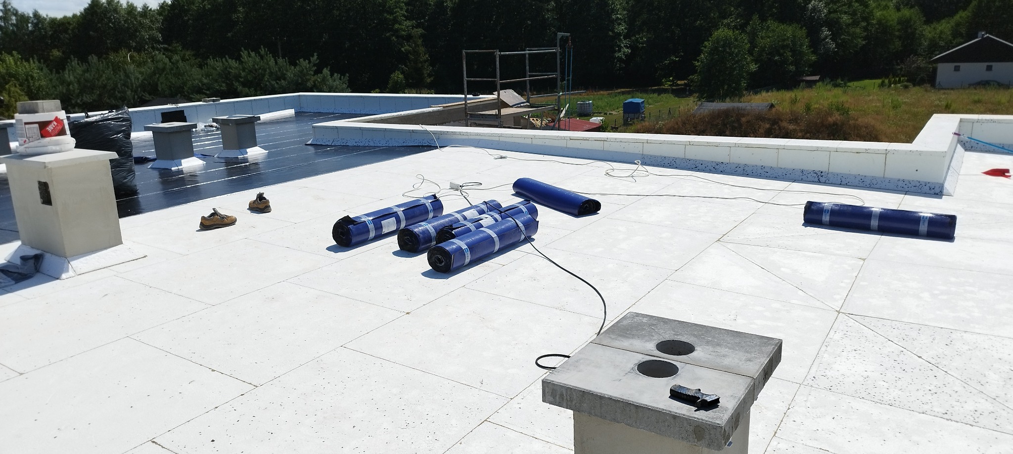 Biały, płaski dach z widocznymi rolkami materiału izolacyjnego w niebieskiej folii, kominami wentylacyjnymi i parą butów roboczych, częściowo pokryty czarną warstwą hydroizolacji.