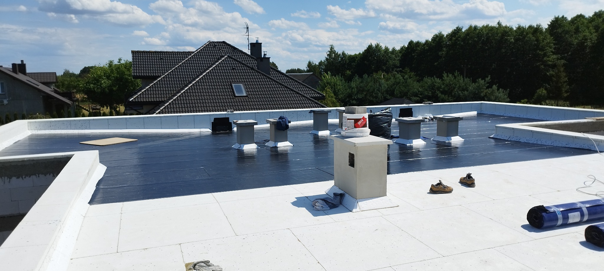 Płaski dach pokryty czarną membraną bitumiczną z wentylacją kominkową, białe obrzeża, w tle budynki mieszkalne i drzewa.