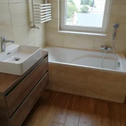 Jasna łazienka po remoncie z umywalką na szafce z szufladami o drewnianym wykończeniu, białą wanną obudowaną płytkami z delikatnym wzorem oraz grzejnikiem drabinkowym.