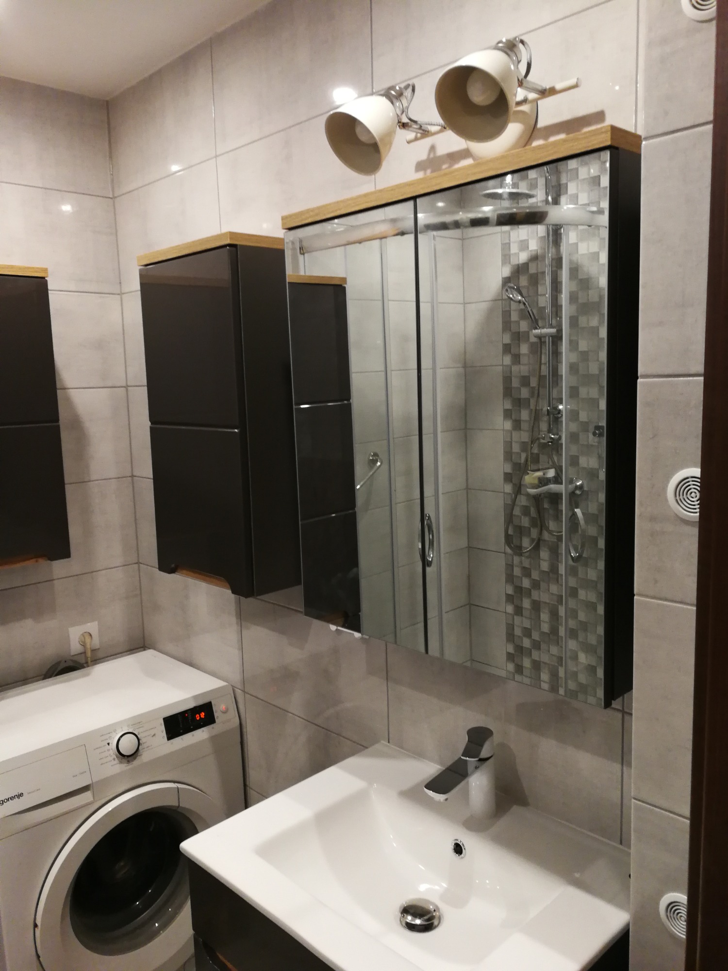 Nowoczesna łazienka z szarymi płytkami, umywalką z szafką, pralką Gorenje i lustrem z oświetleniem, w którym odbija się kabina prysznicowa wyłożona mozaiką.