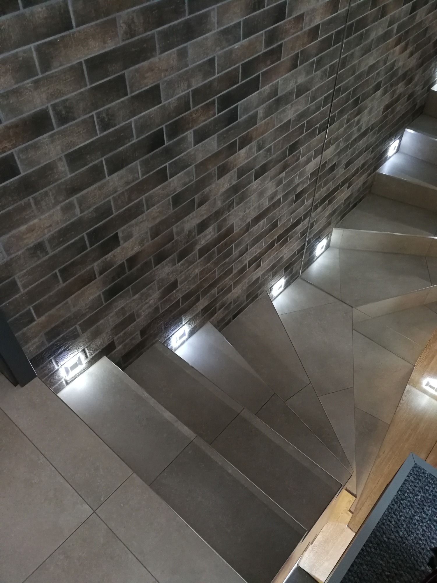 Wykończone schody z szarymi płytkami, podświetlane punktowymi lampami LED w ścianie z cegły klinkierowej w odcieniach brązu i szarości.