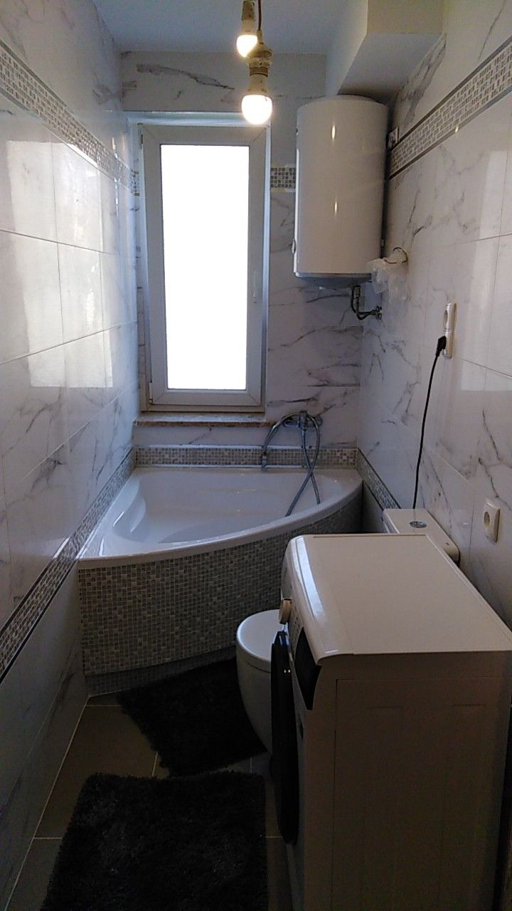 Wnętrze łazienki po remoncie, z narożną wanną obudowaną mozaiką, nową pralką, bojlerem i marmurkowymi płytkami na ścianach. Dwie żarówki zwisają z sufitu.