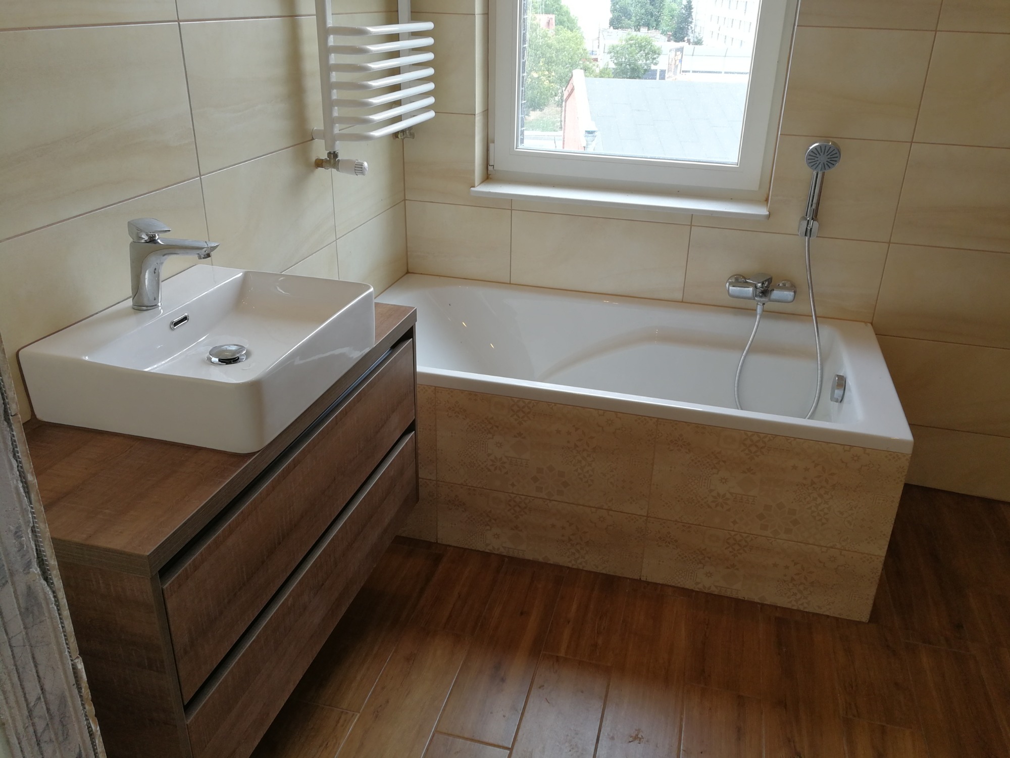 Jasna łazienka po remoncie z umywalką na szafce z szufladami o drewnianym wykończeniu, białą wanną obudowaną płytkami z delikatnym wzorem oraz grzejnikiem drabinkowym.