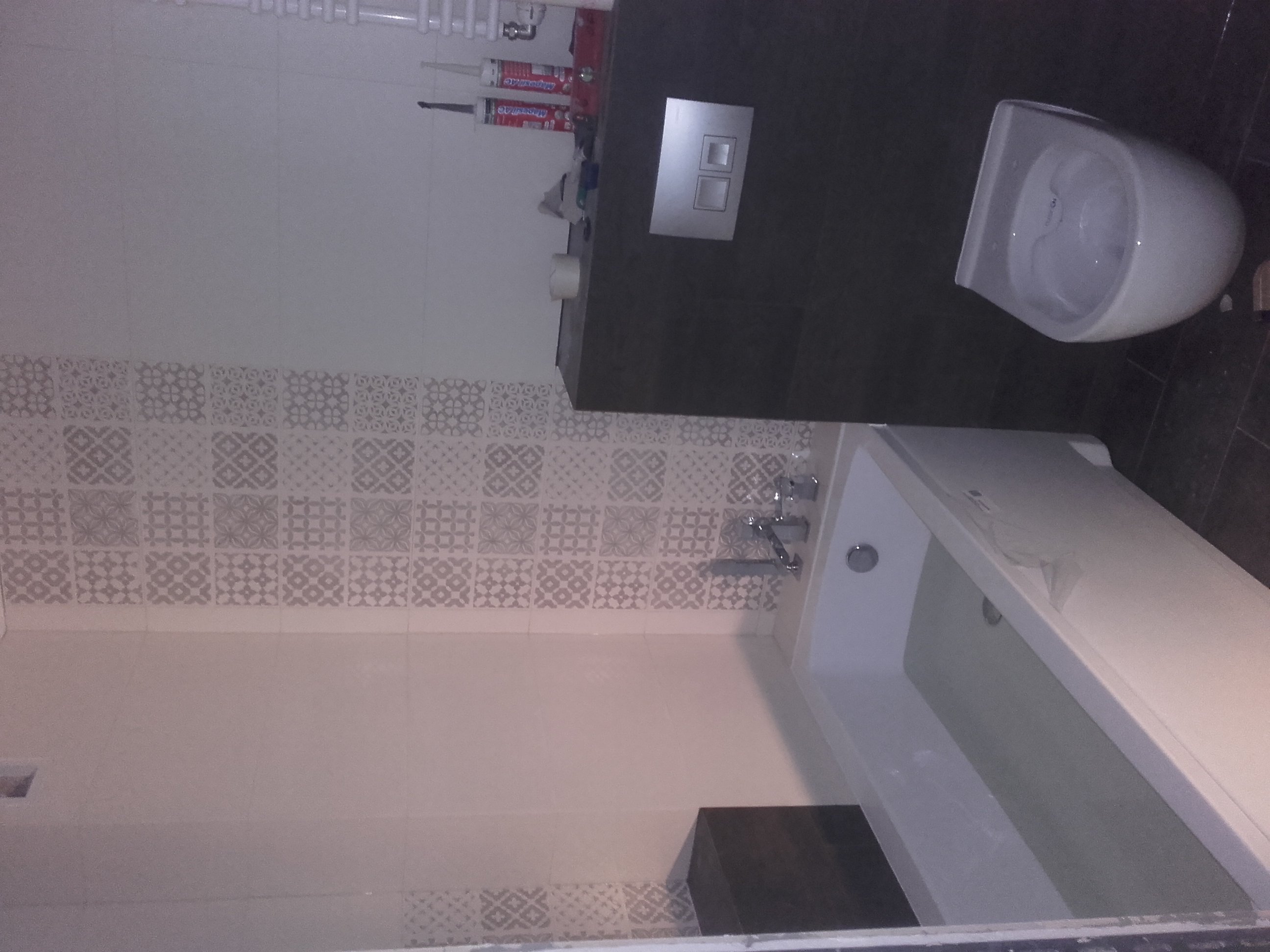 Wykończona łazienka z umywalką, toaletą podwieszaną i ozdobnymi płytkami na ścianie, widoczne tuby z silikonem i włącznik światła.