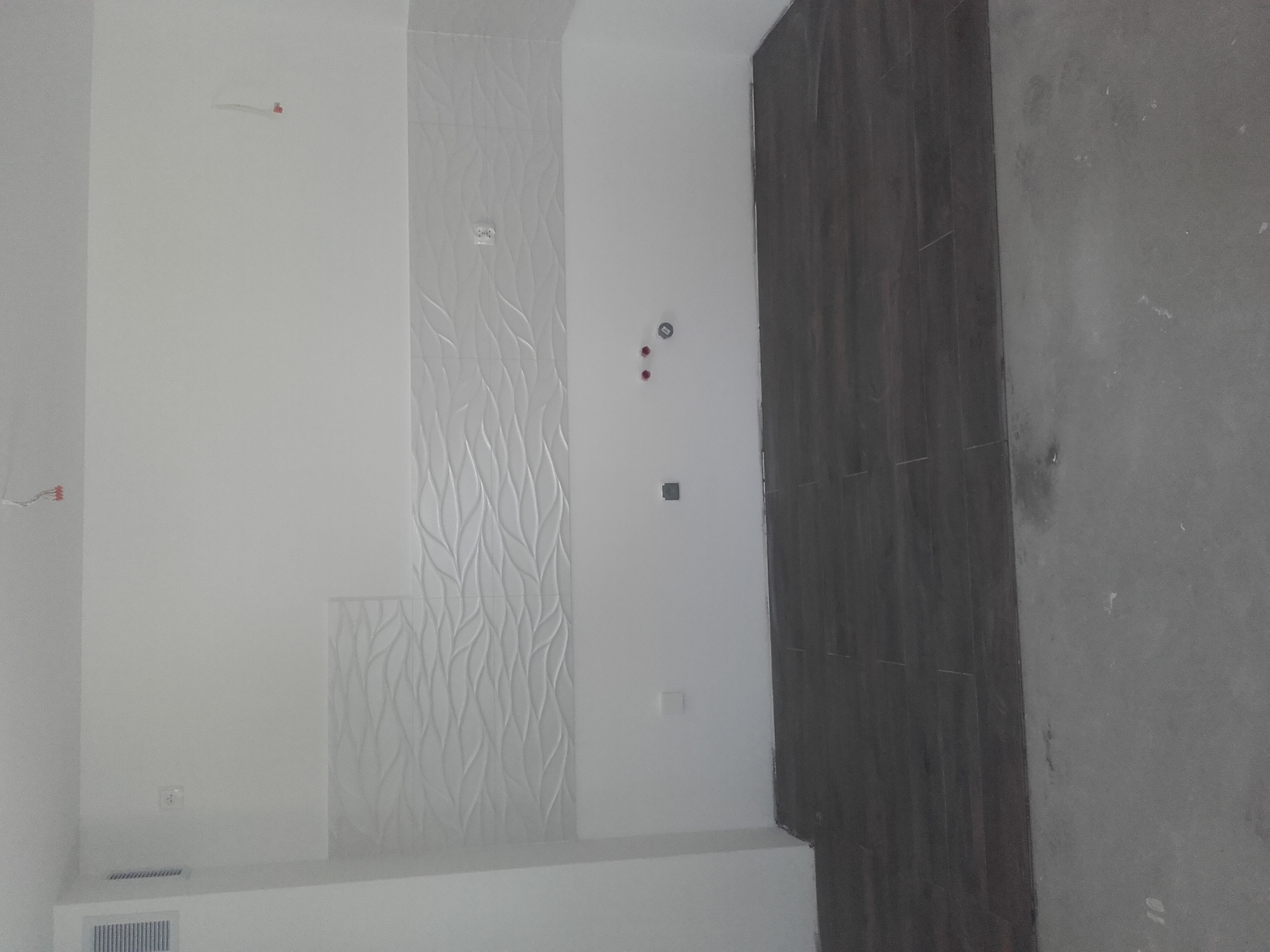 Ściana łazienki wykończona białymi, strukturalnymi płytkami oraz ciemnoszarymi płytkami imitującymi drewno. Widoczne wystające przewody elektryczne i puszki instalacyjne.