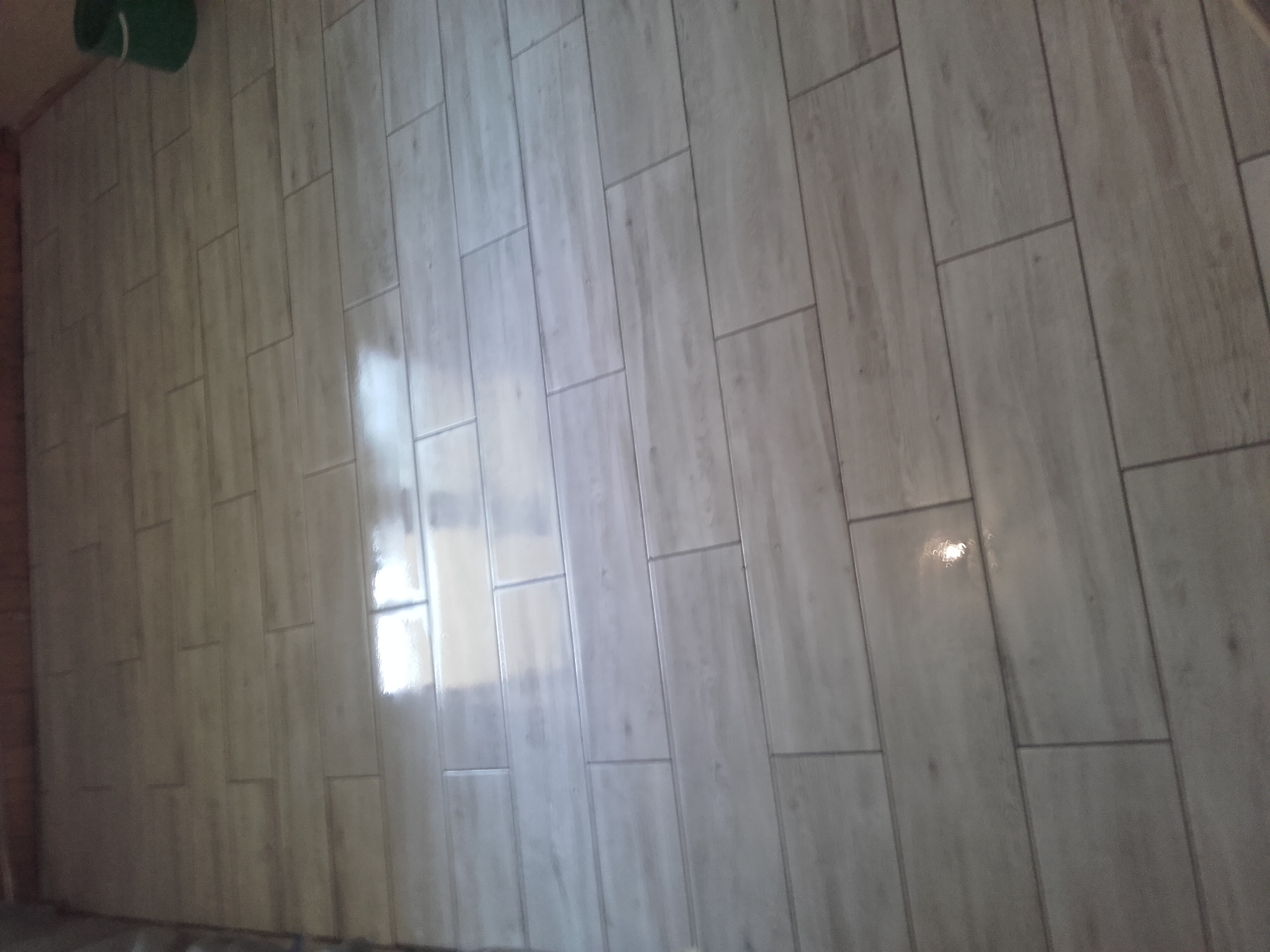 Ułożone podłużne, jasnoszare płytki podłogowe z widocznymi fugami, odbijające światło w górnej części kadru. W lewym górnym rogu widoczny fragment zielonego pojemnika.