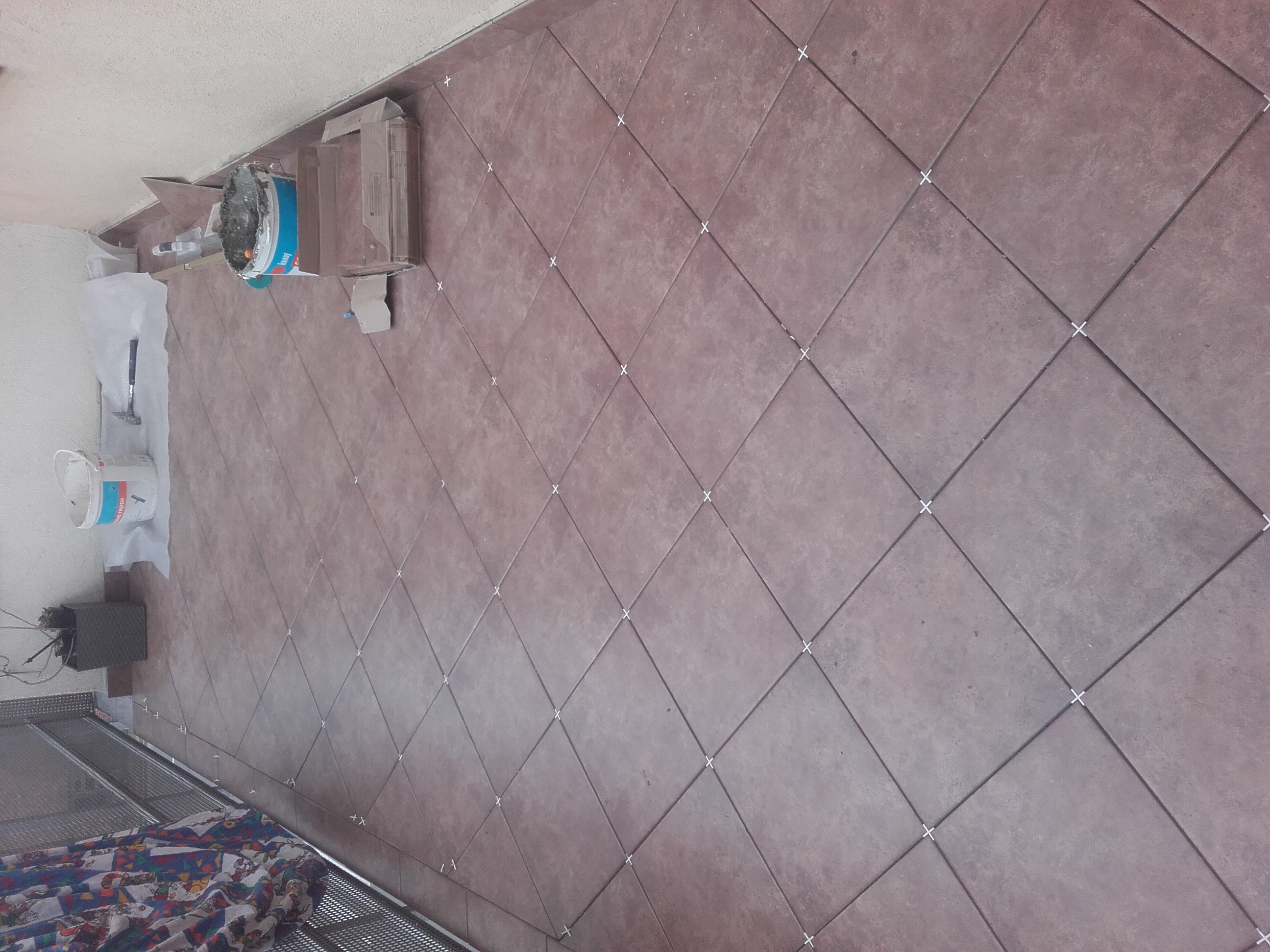 Układanie brązowych płytek podłogowych z białymi krzyżykami dystansowymi, widoczne narzędzia i wiaderka z zaprawą.