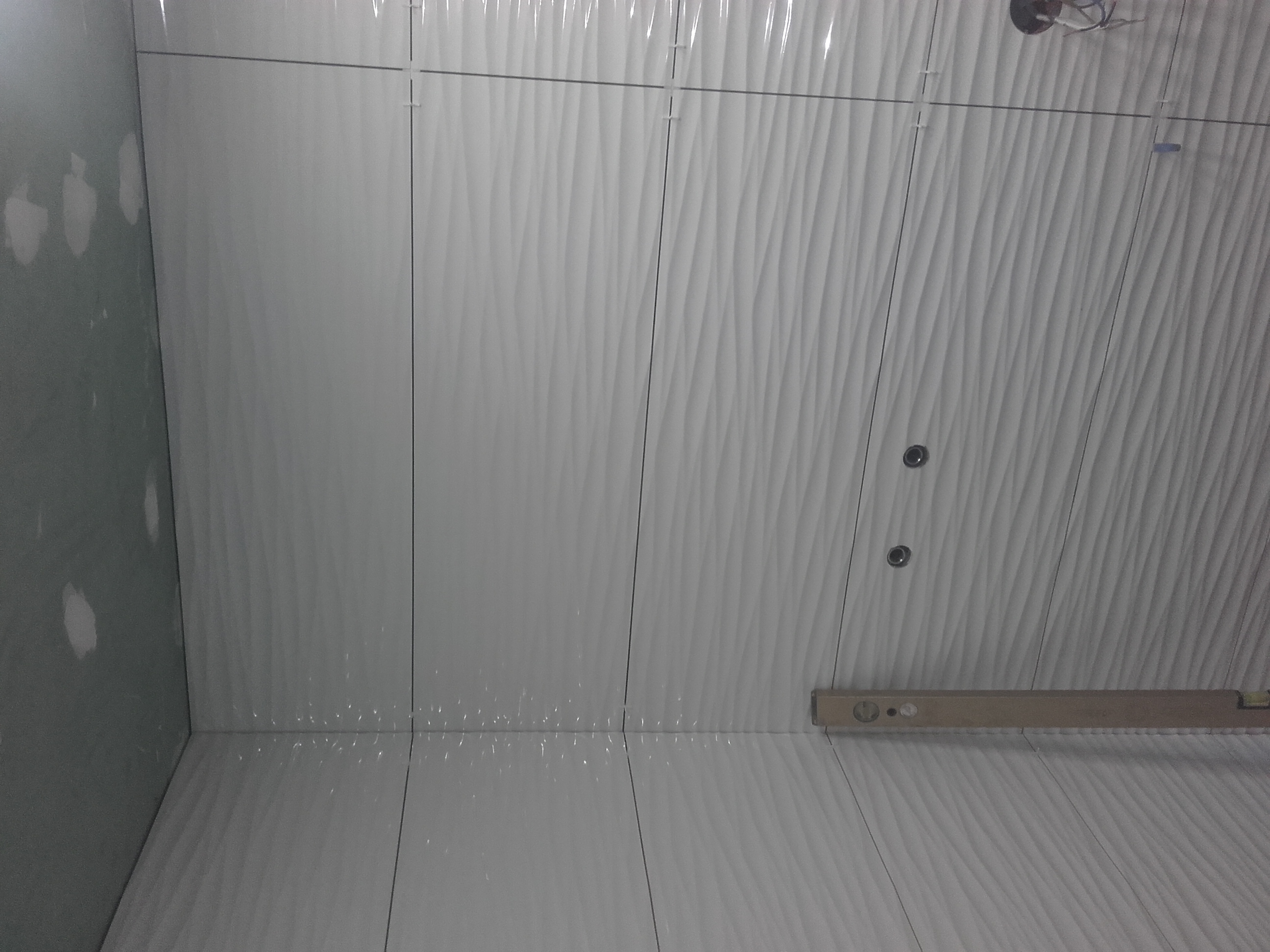 Świeżo położone białe płytki z falistym wzorem na ścianie, z widocznymi krzyżykami dystansowymi i poziomicą przy dolnej krawędzi, oraz fragmentem ściany przygotowanej pod płytki.