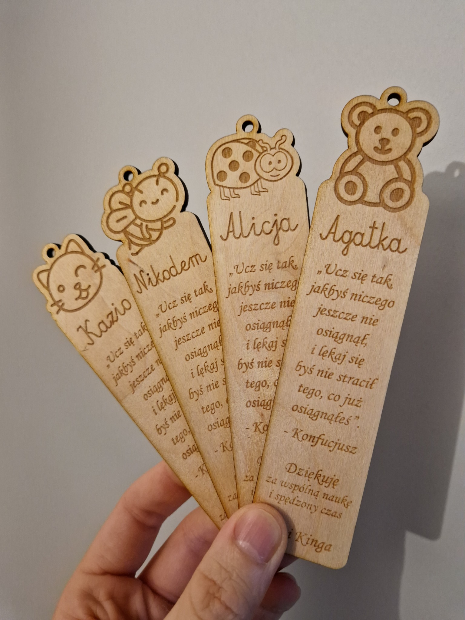 Ręka trzyma wachlarz z czterech drewnianych zakładek do książek, każda z wygrawerowanym imieniem (Kazio, Nikodem, Alicja, Agatka), cytatem Konfucjusza i rysunkiem (kotek, pszczółka, biedronka, miś).