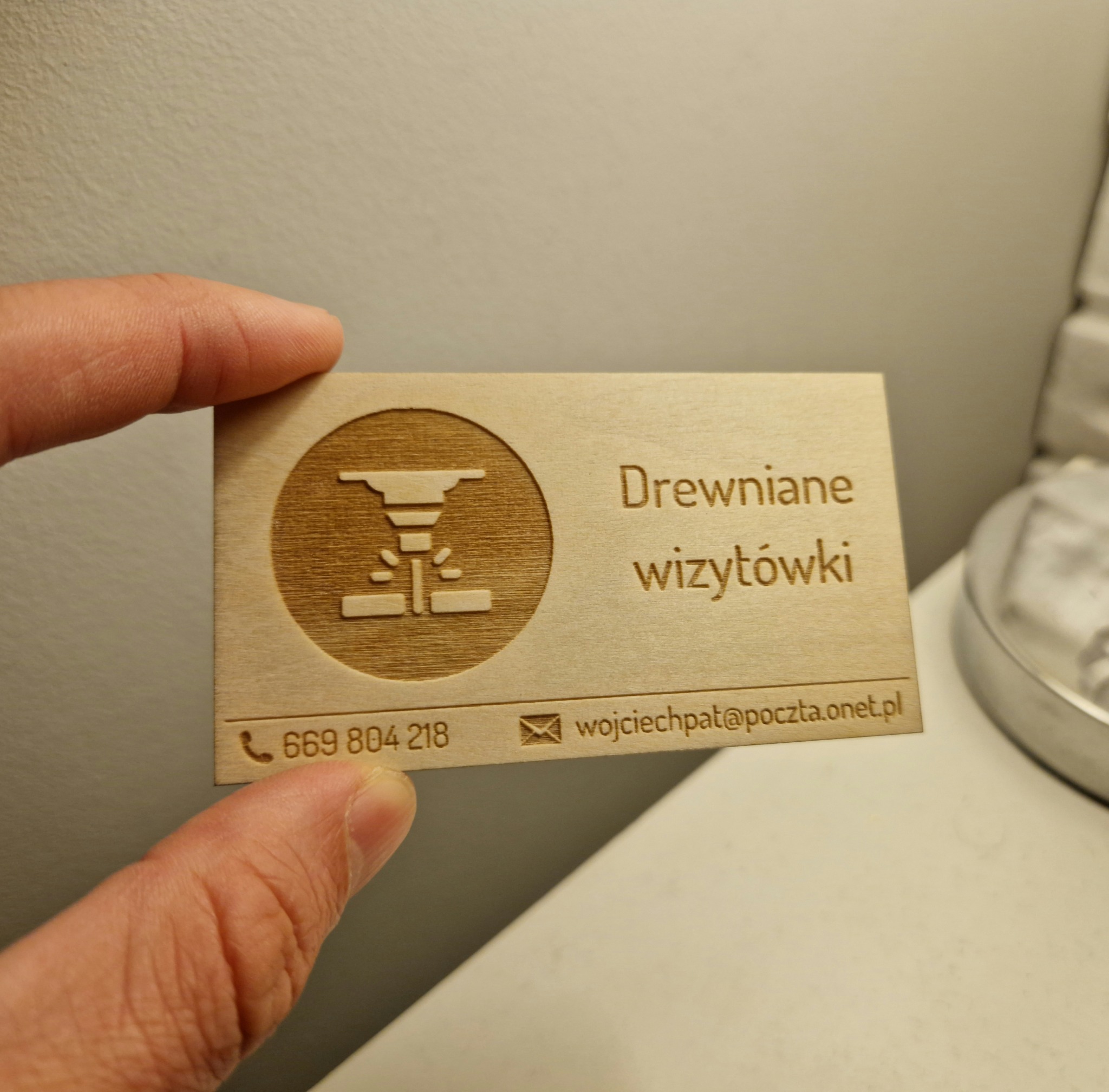 Drewniana wizytówka z wygrawerowanym logo przedstawiającym schemat lasera tnącego, trzymana w palcach na rozmytym tle.