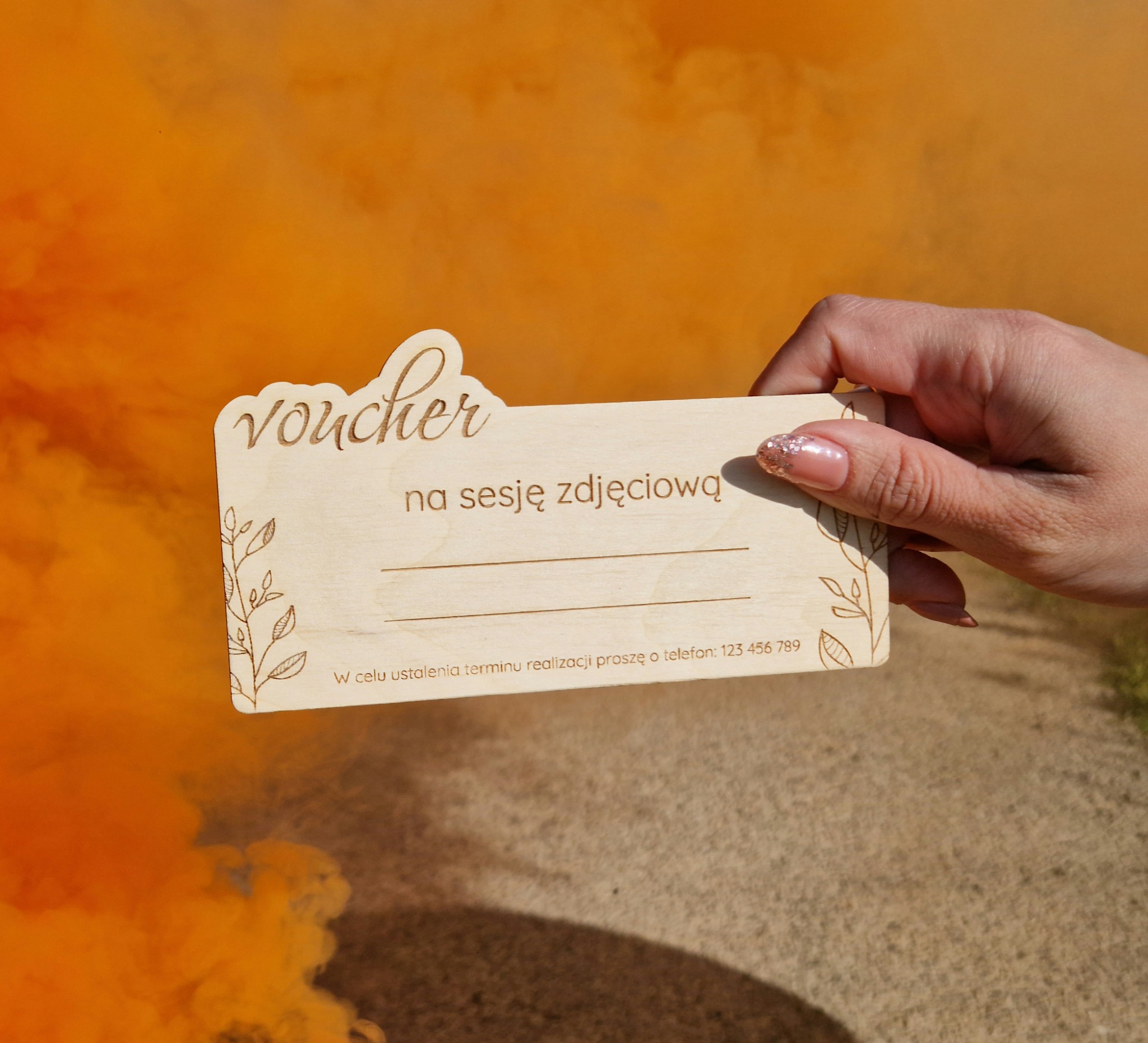 Dłoń trzyma drewniany voucher na sesję zdjęciową z dekoracyjnym napisem i motywem roślinnym, na pomarańczowym, rozmytym tle.