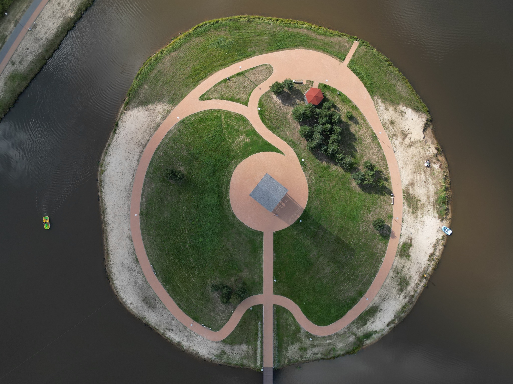 Widok z lotu ptaka na okrągłą wyspę na jeziorze z ścieżkami z kostki brukowej, altaną z czerwonym dachem i zadaszeniem z szarym dachem połączoną mostem z lądem. Na wodzie widoczny rowerek wodny...