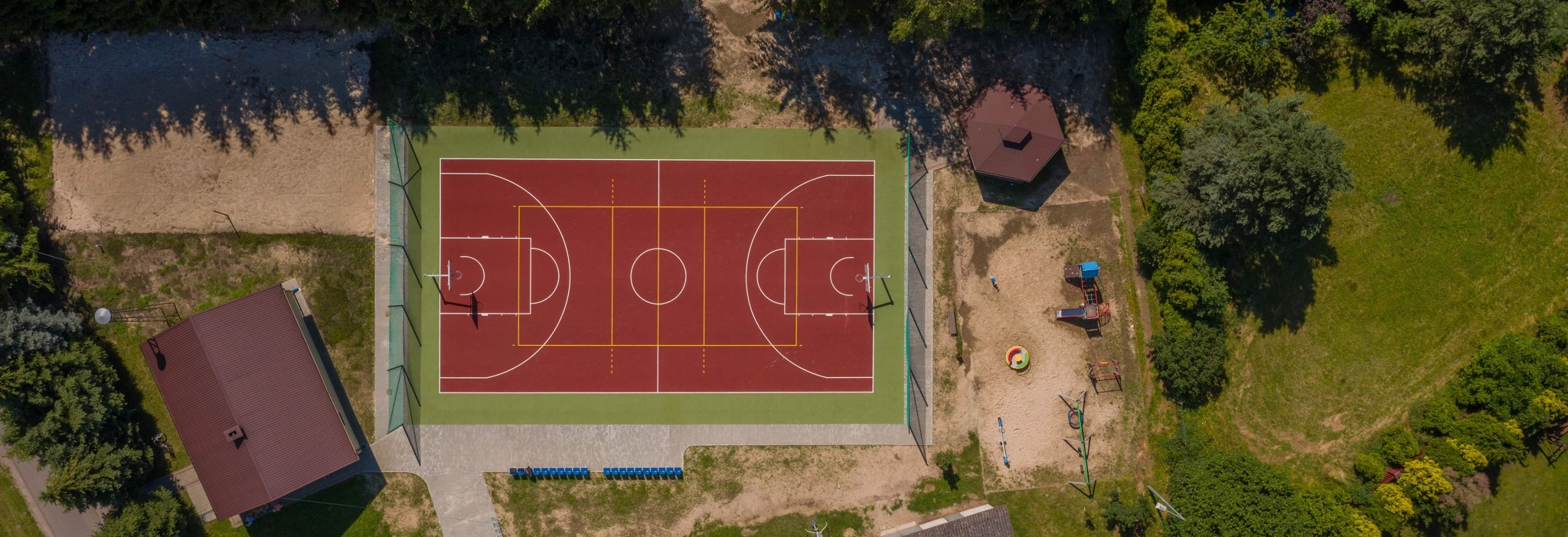 Widok z góry na boisko do koszykówki z czerwoną nawierzchnią i zielonym obramowaniem, obok plac zabaw dla dzieci z piaskiem i domkiem z brązowym dachem.