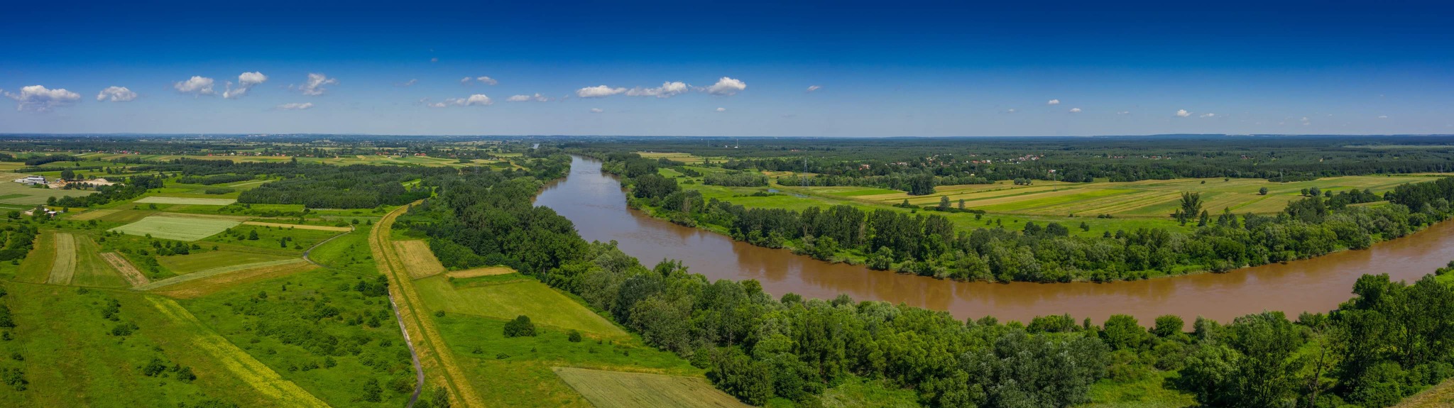 Panoramiczny widok z lotu ptaka na rozległą rzekę wijącą się przez zielone pola i lasy pod błękitnym niebem z lekkimi obłokami.