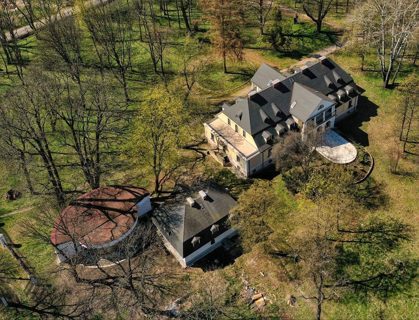 Zdjęcie z lotu ptaka przedstawia rozległą posiadłość z budynkiem głównym w stylu klasycystycznym z ciemnym dachem i żółtą elewacją oraz okrągłą przybudówką z czerwonym dachem, otoczoną drzewami...