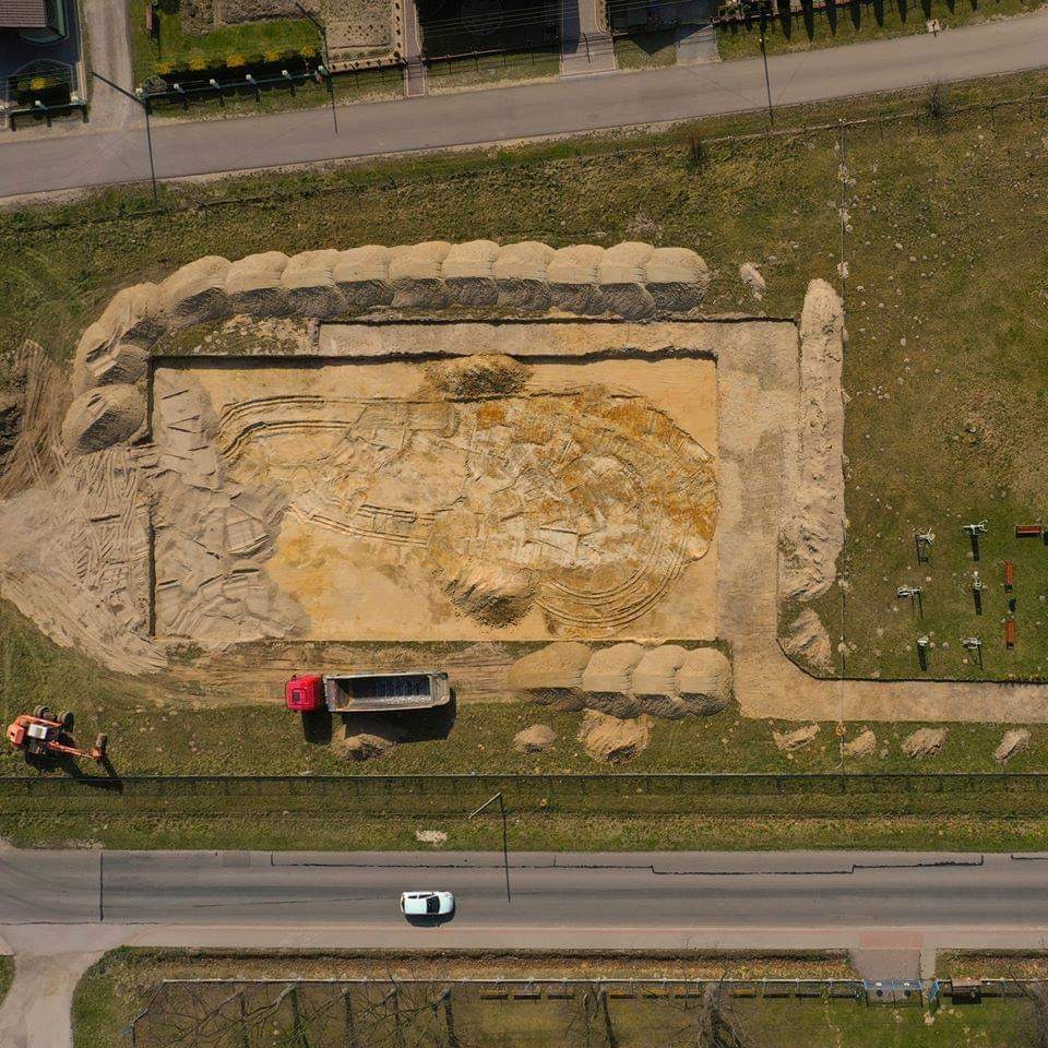 Zdjęcie z drona przedstawia plac budowy z wykopem w kształcie okręgu, otoczonym wałem z piasku, obok stoją maszyny budowlane i ciężarówka.