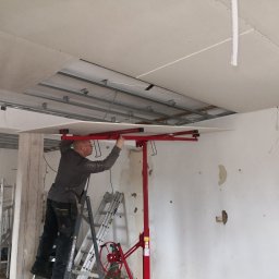 Pan Krzysztof Rogalski - Pracownik montuje płytę gipsowo-kartonową do sufitu za pomocą podnośnika, widoczne metalowe profile konstrukcyjne i fragmenty instalacji elektrycznej.